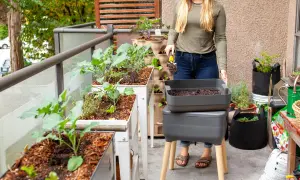 Une femme prépare son compost d'appartement sur un balcon verdoyant, avec des bacs de culture et un lombricomposteur.