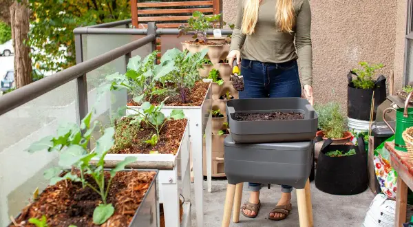 Une femme prépare son compost d'appartement sur un balcon verdoyant, avec des bacs de culture et un lombricomposteur.