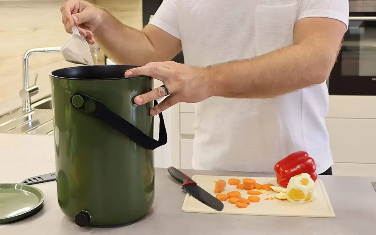 Un homme ajoute un sachet dans un seau vert, un ustensile pratique pour le compost appartement. Des carottes coupées et un poivron rouge sont sur une planche à découper.