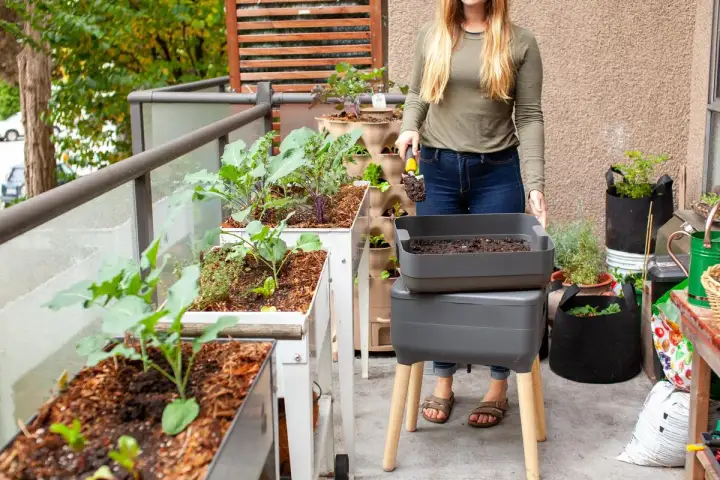 Une femme prépare son compost d'appartement sur un balcon verdoyant, avec des bacs de culture et un lombricomposteur.